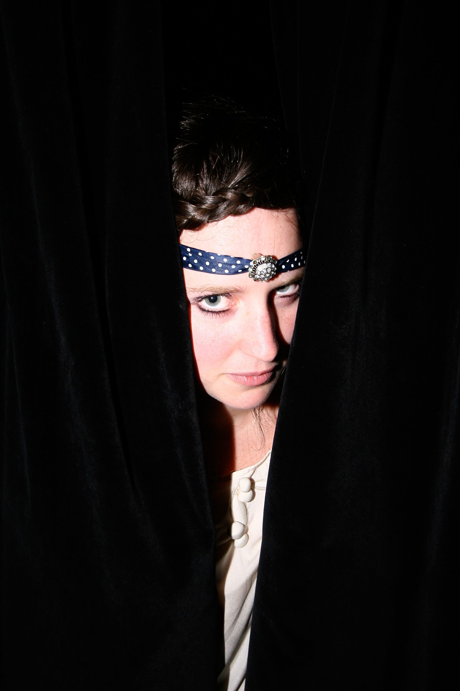 A woman in a polka-dot headband peers out from behind a black curtain.